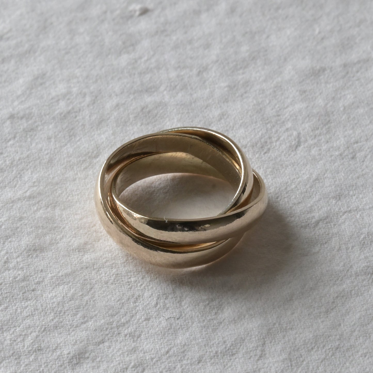 A yellow gold trinity ring with three interlinked bands, displayed on a textured background.