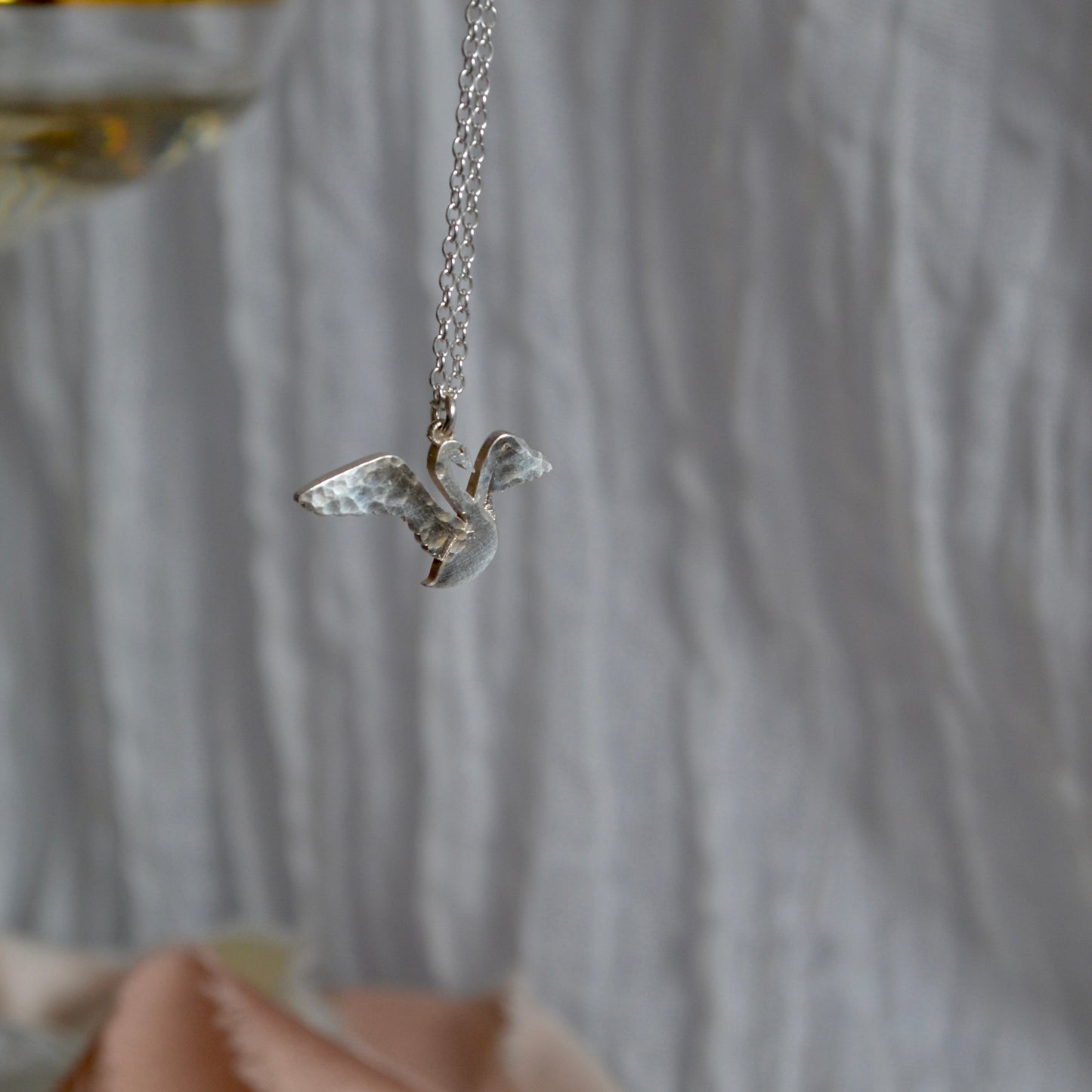 Silver bird-shaped pendant necklace on a blurred background with a glass of wine.