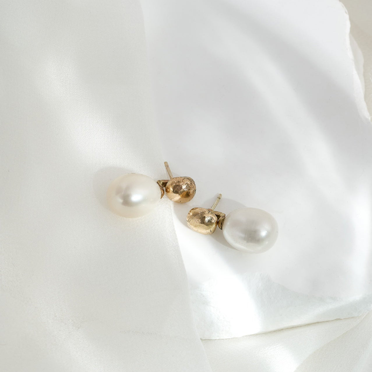 A close-up image of a gold pearl drop stud earring on a white textured surface.
