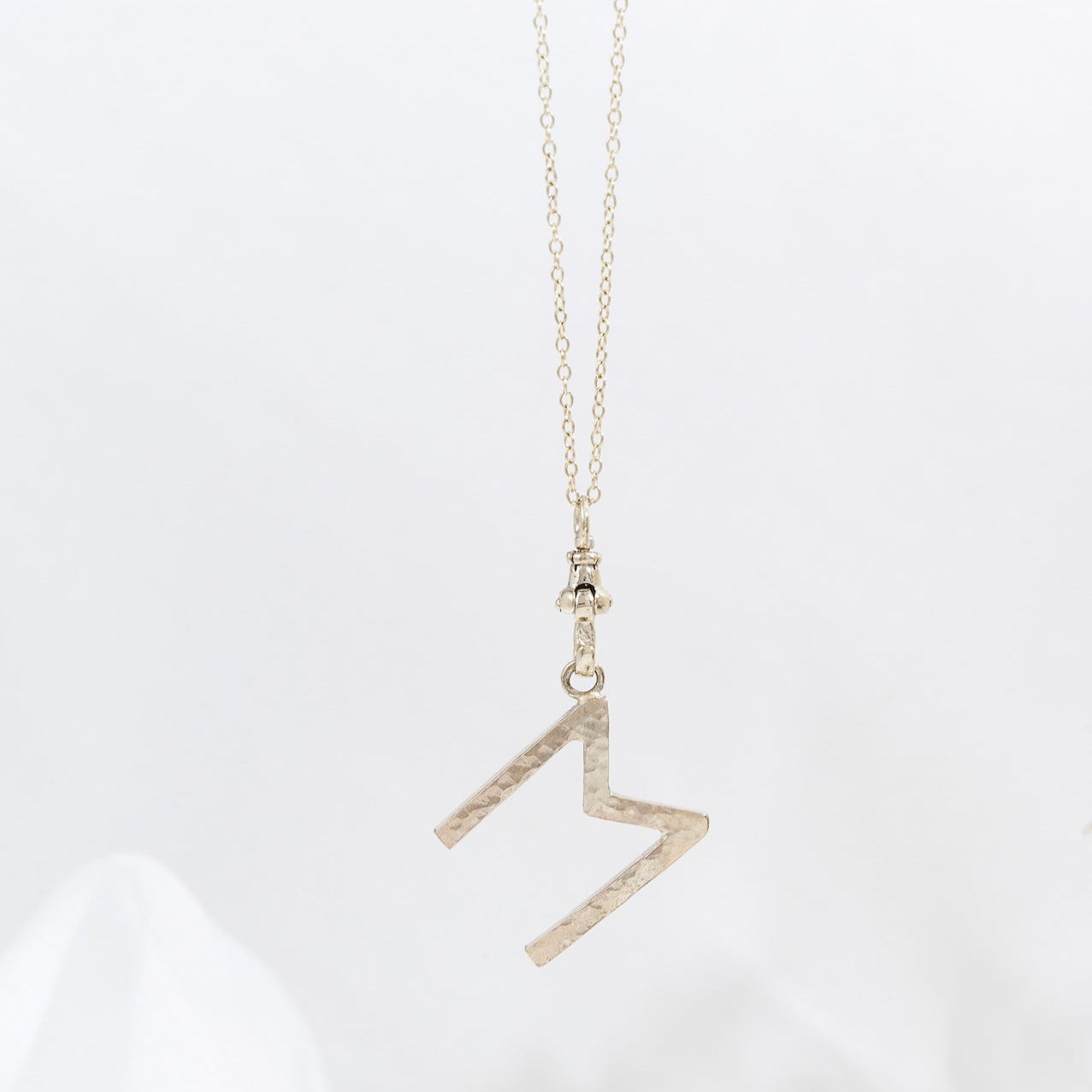A gold letter pendant necklace on a white background, with the pendant in the shape of the letter 'M' and the necklace chain visible.