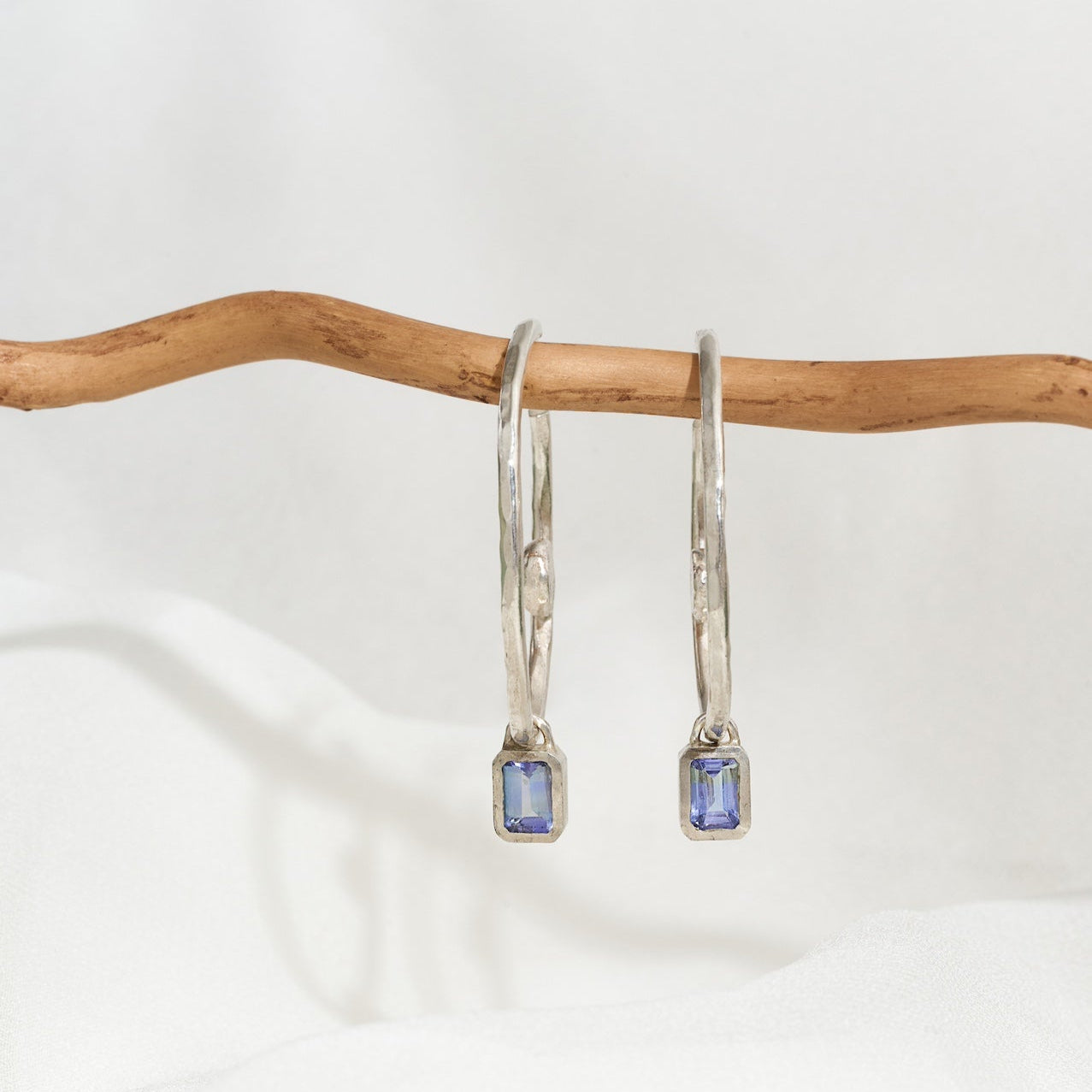 A pair of sterling silver hoop earrings with blue tanzanite gemstones, displayed against a white background.