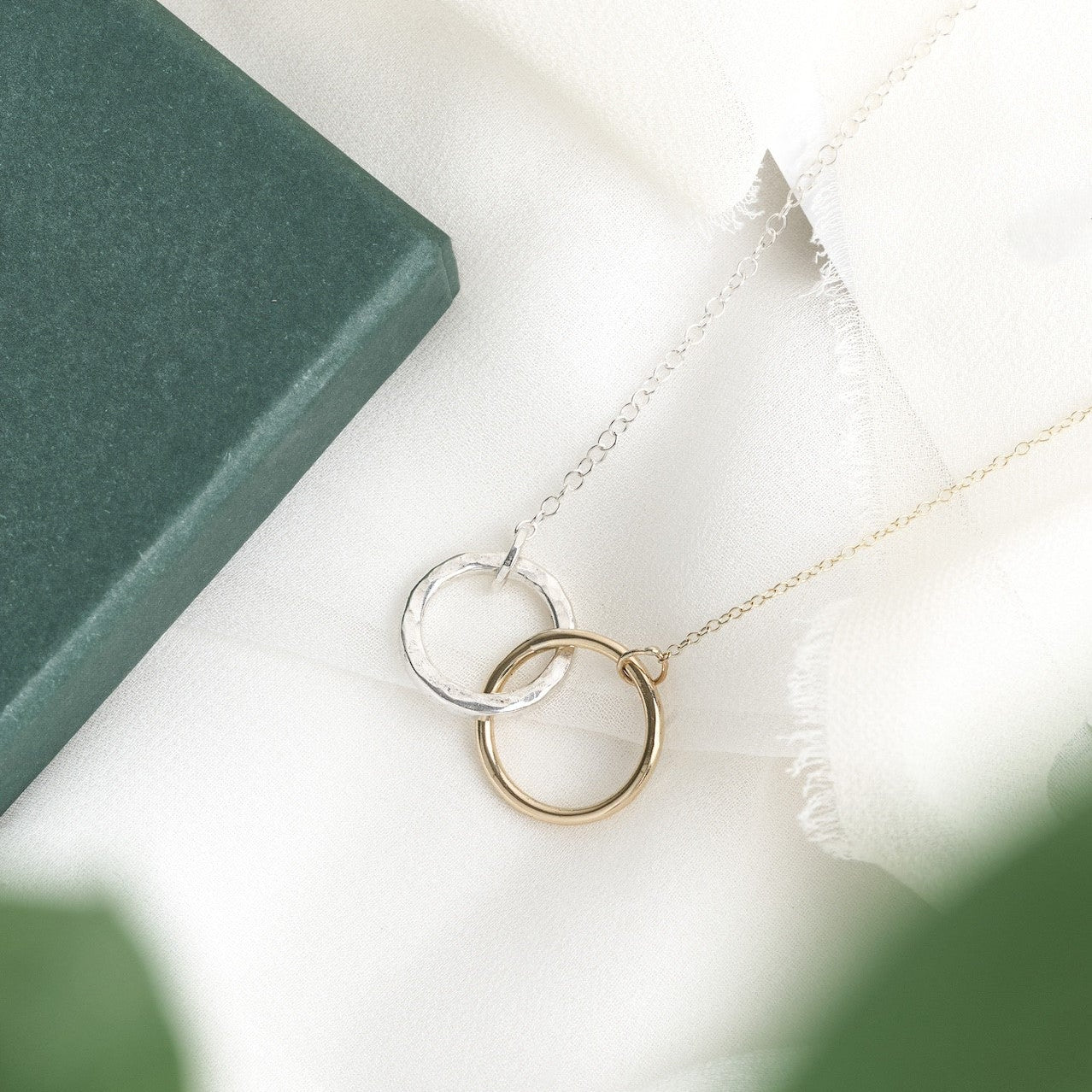 A gold and silver interlocking infinity necklace placed on a white surface next to a green book and a white flower, with a blurred green leaf in the foreground.