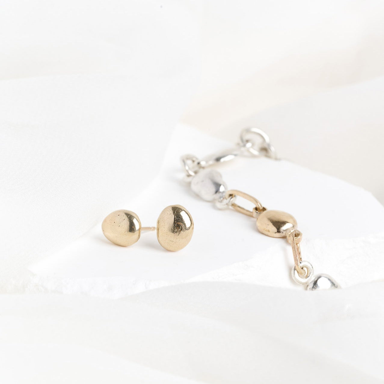 A silver pebble bracelet with a gold bead at the center, alongside a pair of gold river pebble stud earrings, placed on a white fabric surface.