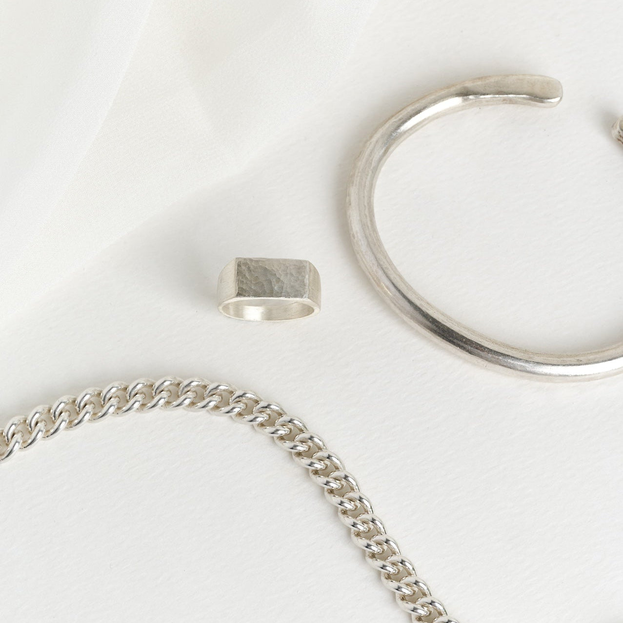 A set of silver jewelry including a bracelet with a lobster clasp, a chunky silver cuff, and a silver rectangle dappled signet, displayed on a white fabric surface.