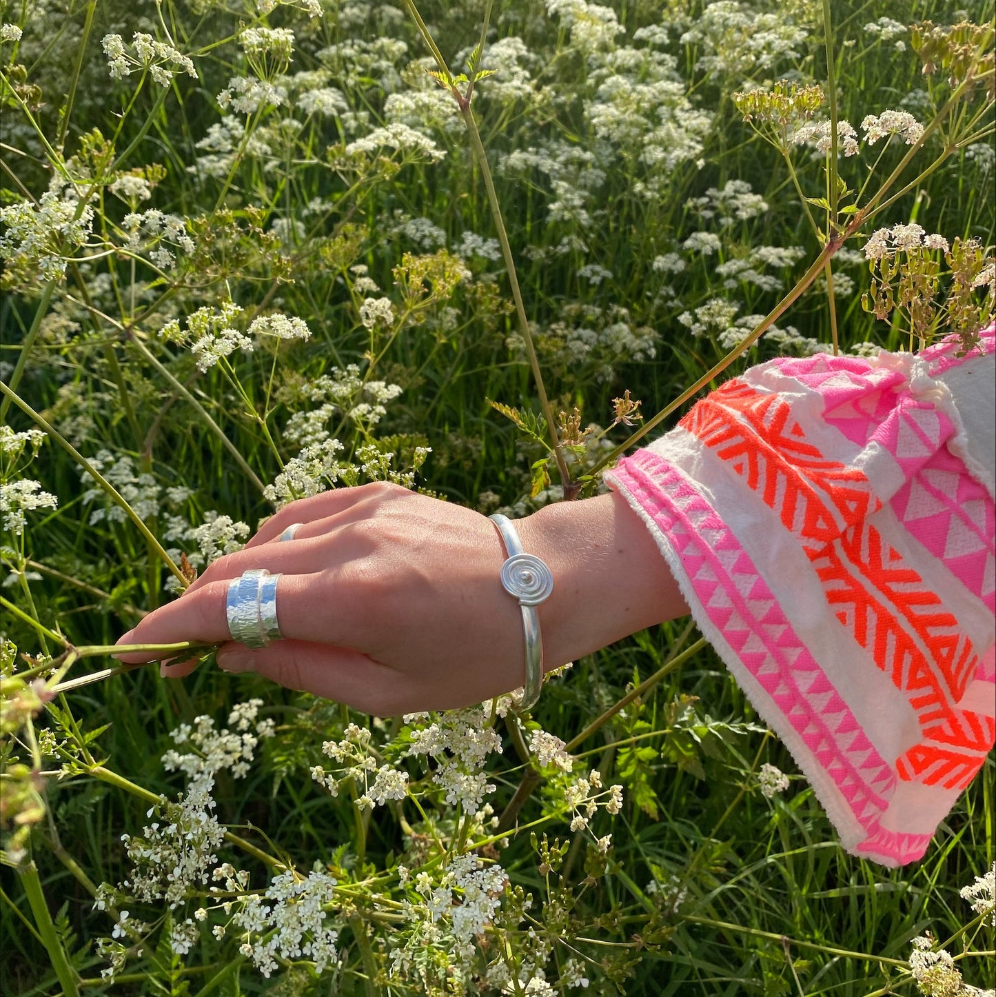 A person's forearm showcasing a silver bangle with a spiral design, worn on a wrist with a lush green field in the background.
