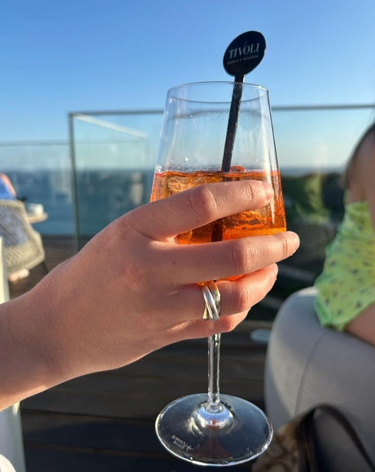 A sterling silver and 9ct yellow gold trinity ring  worn on a females hand, holding an aperol spritz while on holiday enjoying the sunshine. 