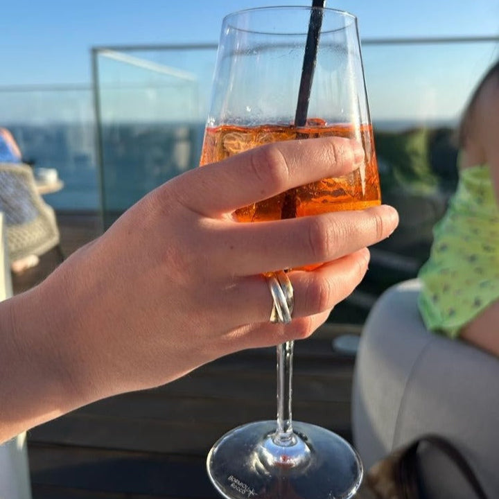 A sterling silver and 9ct yellow gold trinity ring  worn on a females hand, holding an aperol spritz while on holiday enjoying the sunshine. 