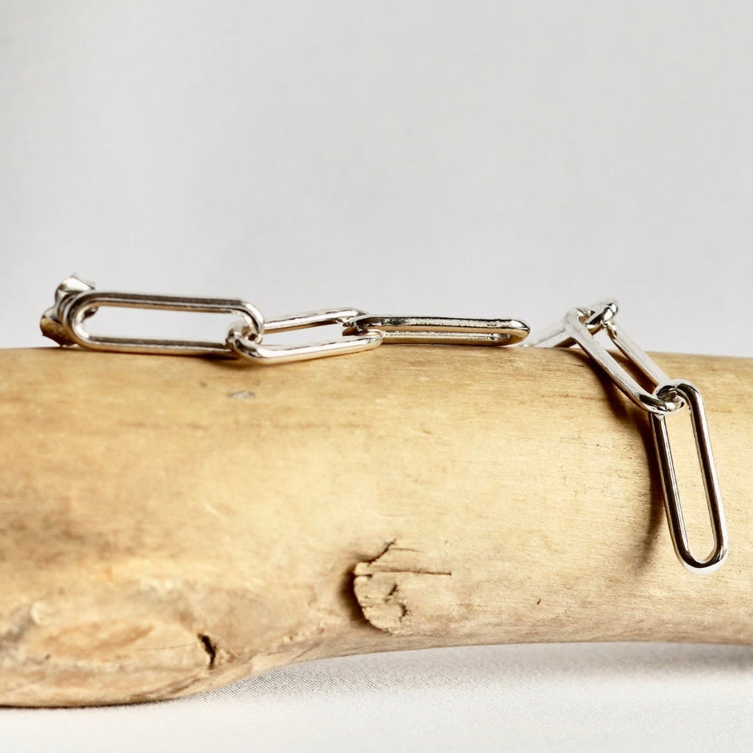 A pair of silver paperclip earrings displayed on a light beige driftwood log against a neutral light gray background.