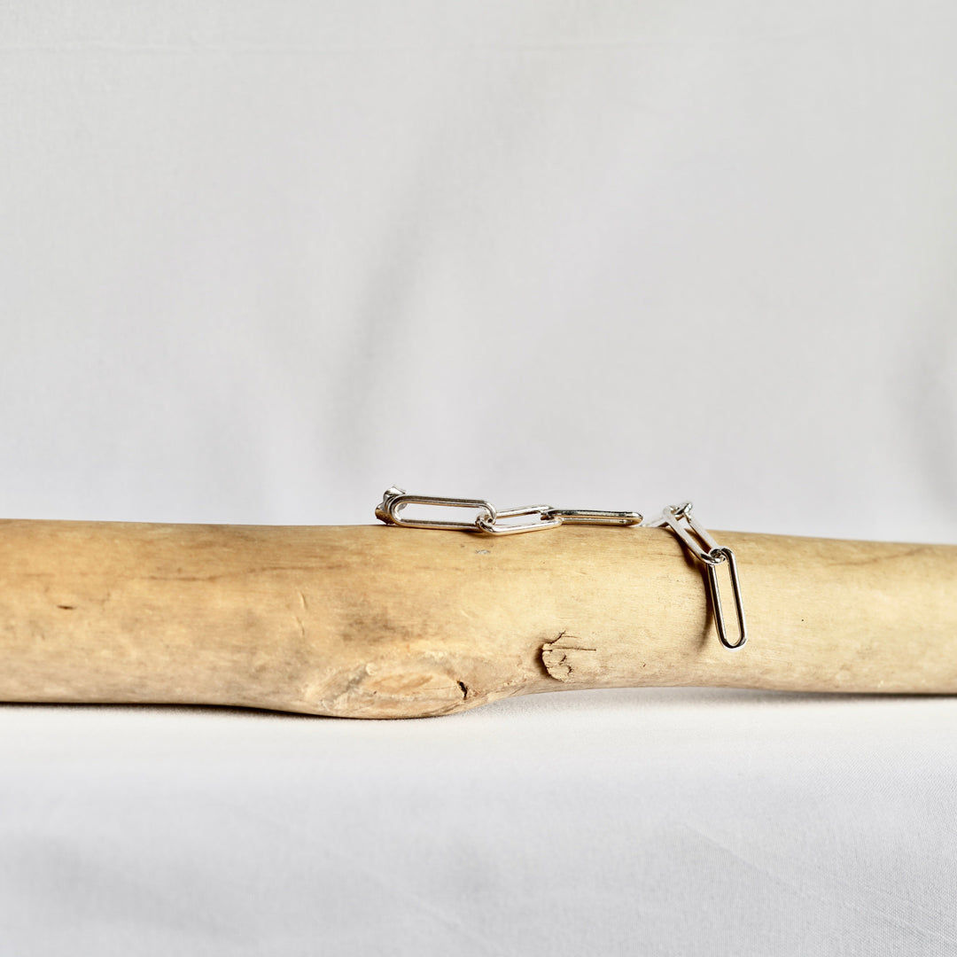 A pair of silver paperclip earrings displayed on a light tan driftwood log against a white fabric background.