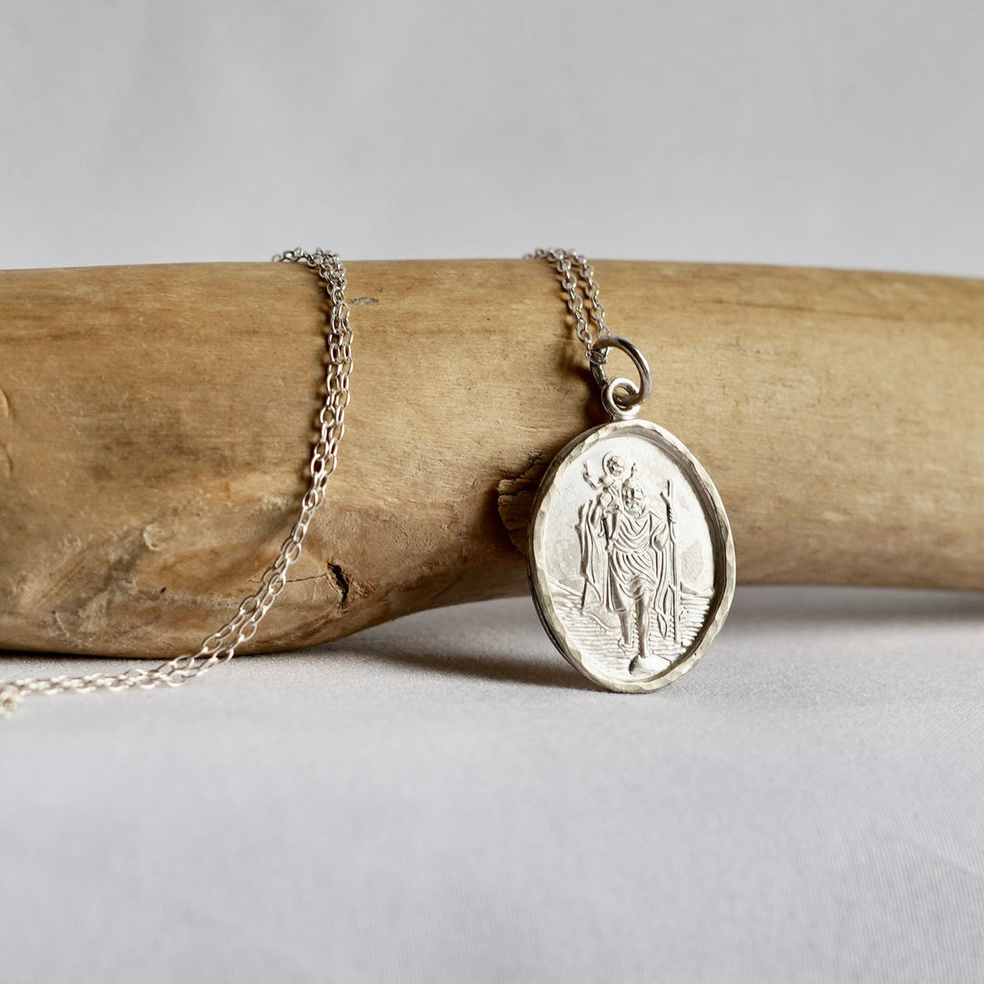 A silver oval pendant necklace featuring an embossed religious figure, draped over a light brown driftwood branch against a neutral gray background.