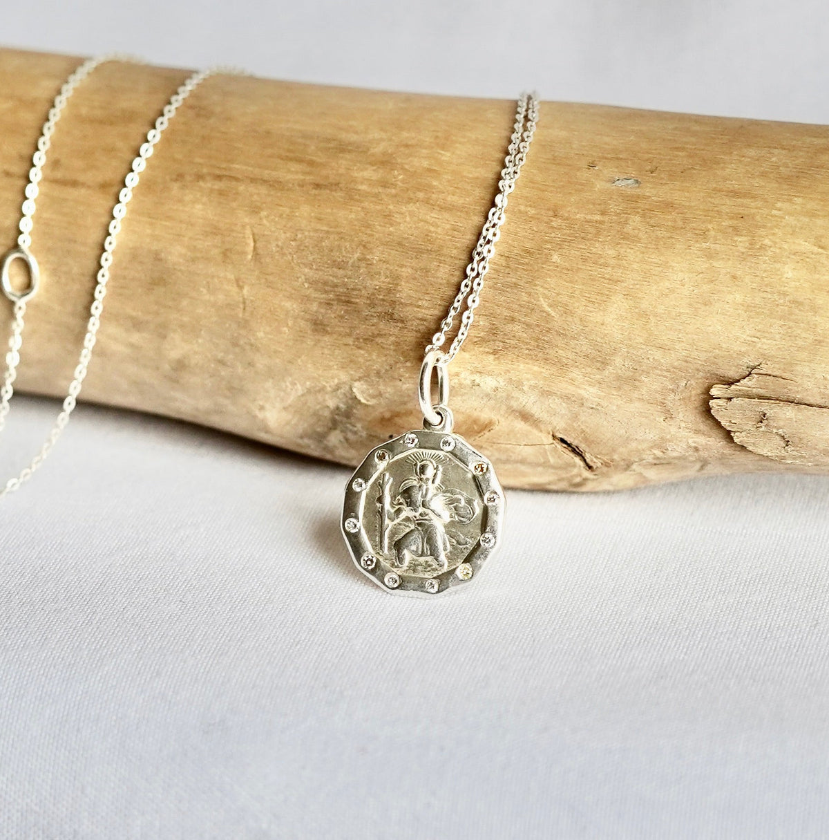 A silver chain necklace with a round medallion pendant featuring a religious figure, displayed on a light wood branch against a white fabric background.