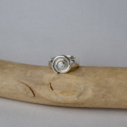 Silver ring with a circular design on a piece of driftwood against a gray background