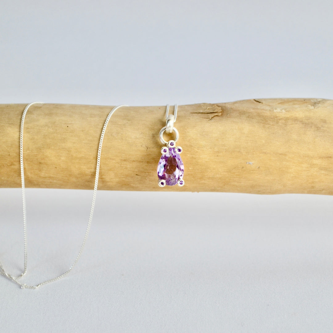 Necklace with a purple teardrop pendant on a wooden stick against a light gray background