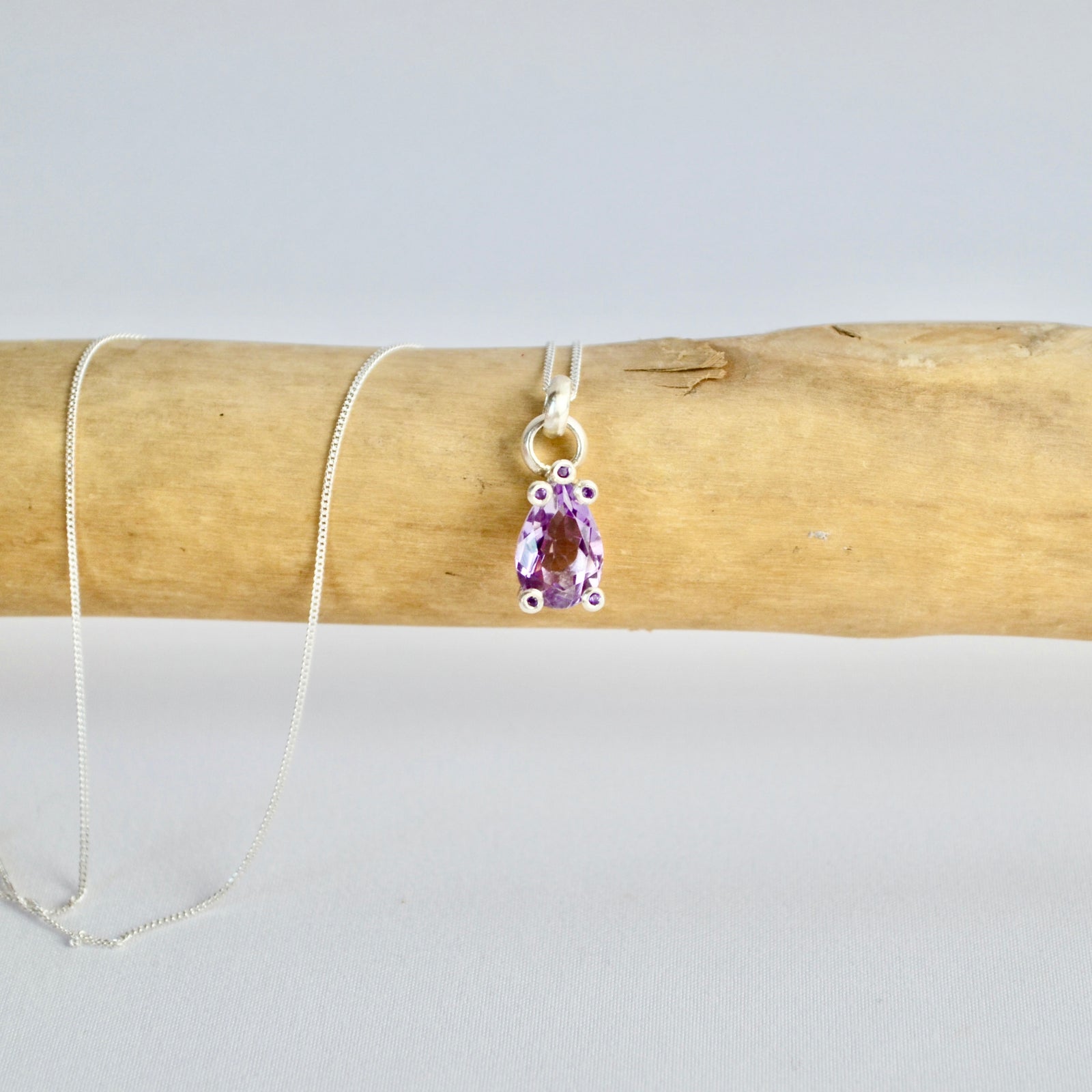 Necklace with a purple teardrop pendant on a wooden stick against a light gray background