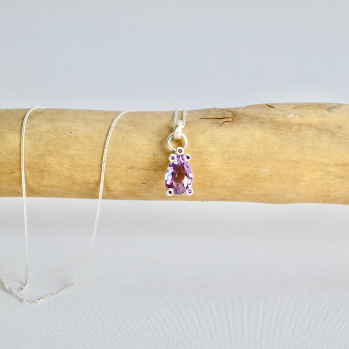 Necklace with a purple teardrop pendant on a wooden stick against a light gray background