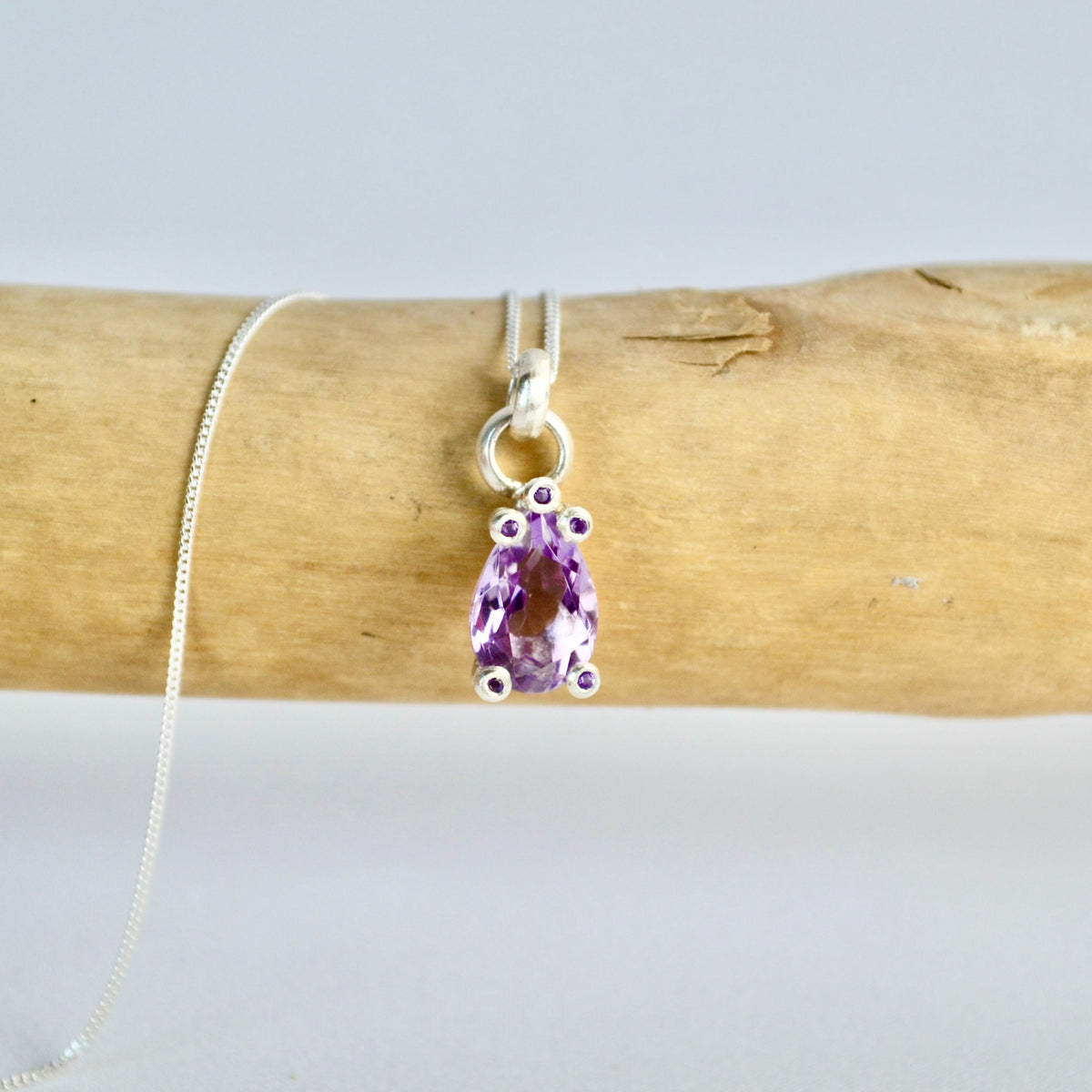 Amethyst pendant on a silver chain draped over wooden logs with a white background