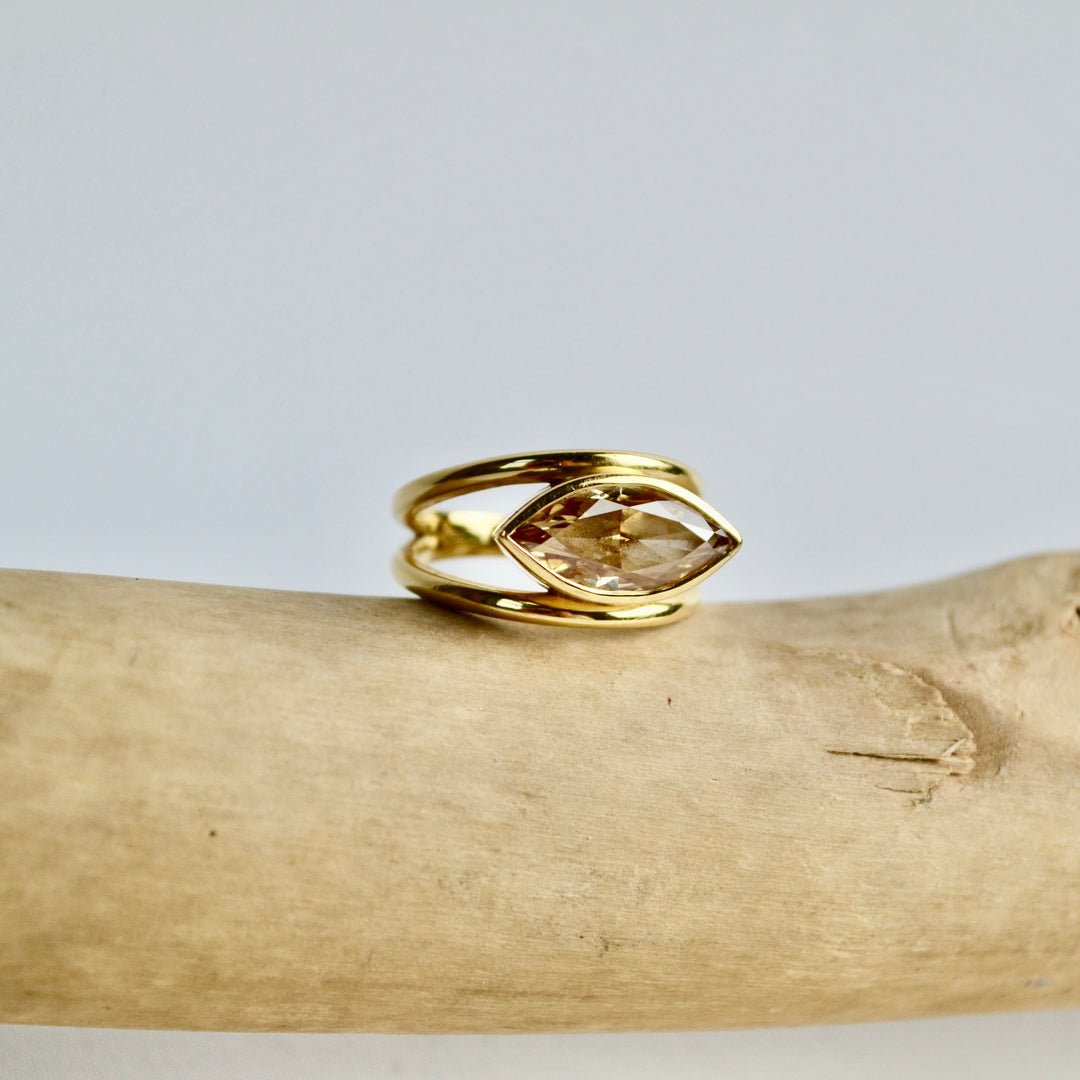 Gold ring with a clear gemstone on a wooden branch against a light gray background