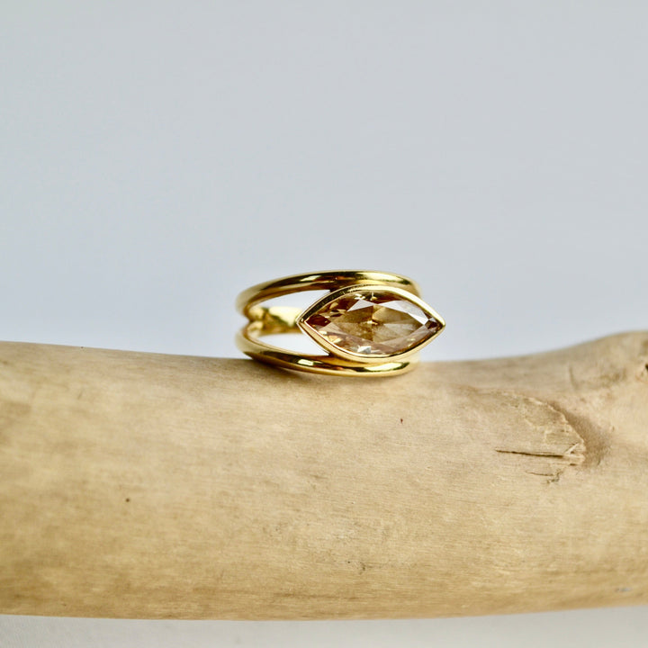 Gold ring with a clear gemstone on a wooden branch against a light gray background