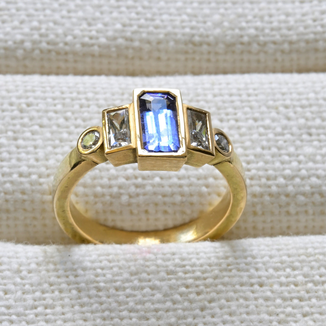 A gold band ring with a central tanzanite gemstone and two adjacent diamonds, placed on a white textured surface.