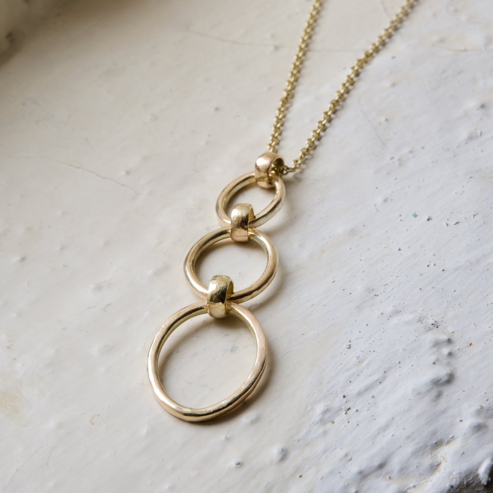 A gold necklace with a solid pendant featuring ascending ovals, attached to a yellow gold curb chain, displayed against a white textured background.