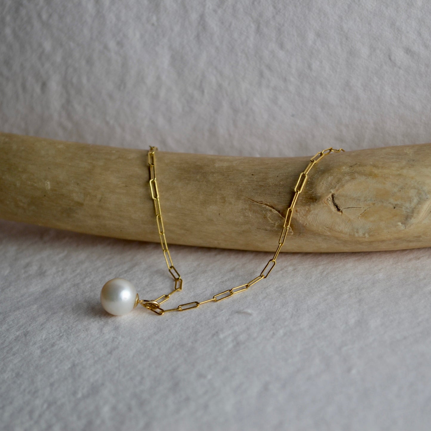 Gold chain bracelet with a pearl on a wooden stick against a textured white background