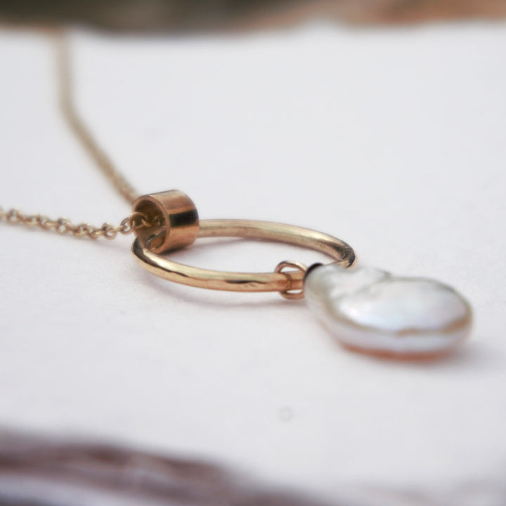 A gold-colored necklace featuring a pear-shaped white pearl pendant, attached to a gold hoop with a curb chain.