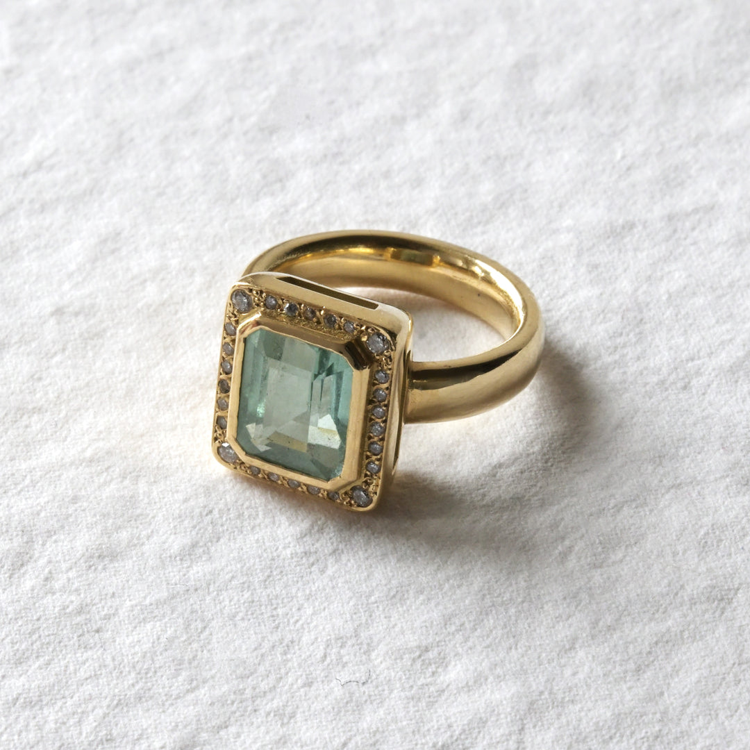An 18ct yellow gold engagement ring with a mint green tourmaline stone set centrally, surrounded by white brilliant diamonds, on a textured white background.