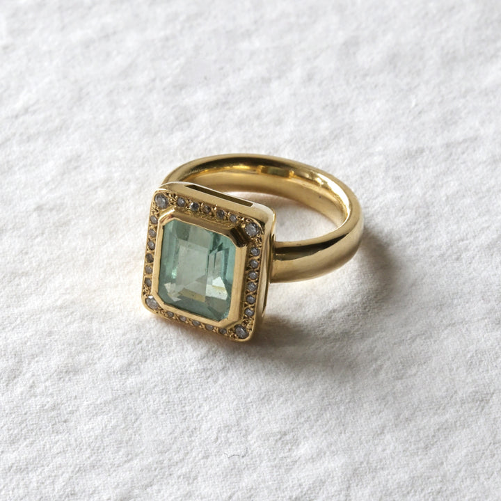 An 18ct yellow gold engagement ring with a mint green tourmaline stone set centrally, surrounded by white brilliant diamonds, on a textured white background.