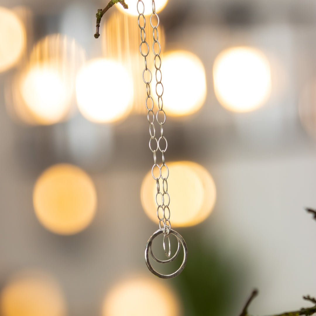 A sterling silver necklace with three connected circular pendants on a teardrop chain, displayed against a blurred background of lights.