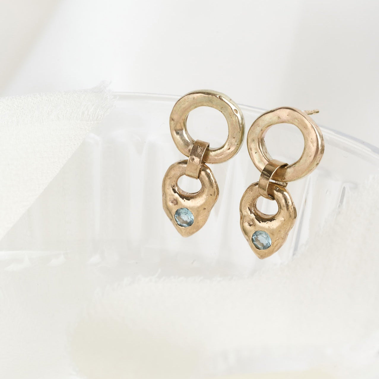 A pair of gold and aquamarine earrings with a teardrop-shaped pale blue aquamarine drop and a chunky gold circle at the top, displayed on a reflective surface.