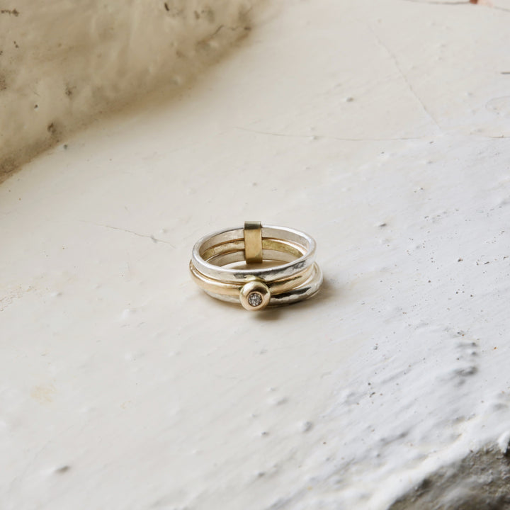 A ring with two silver bands and one gold band, featuring a white brilliant natural diamond set in a gold molten ball at its centre, placed on a textured white surface.
