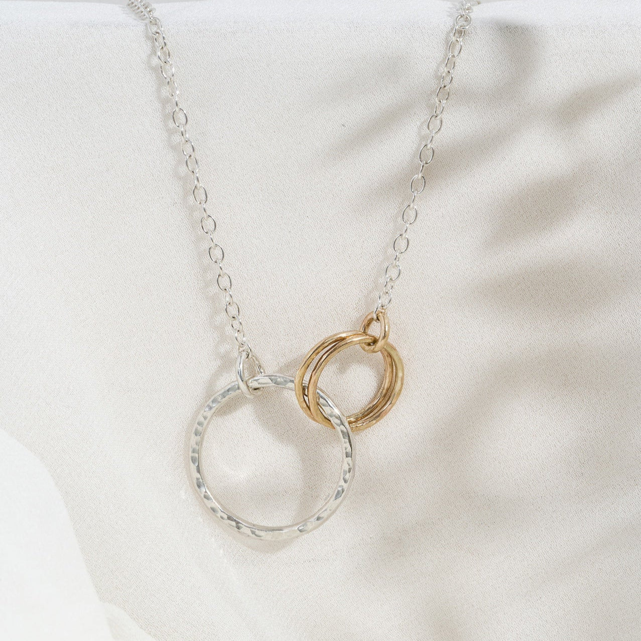 A silver necklace featuring two gold circles and a gold chain, displayed on a white fabric surface.