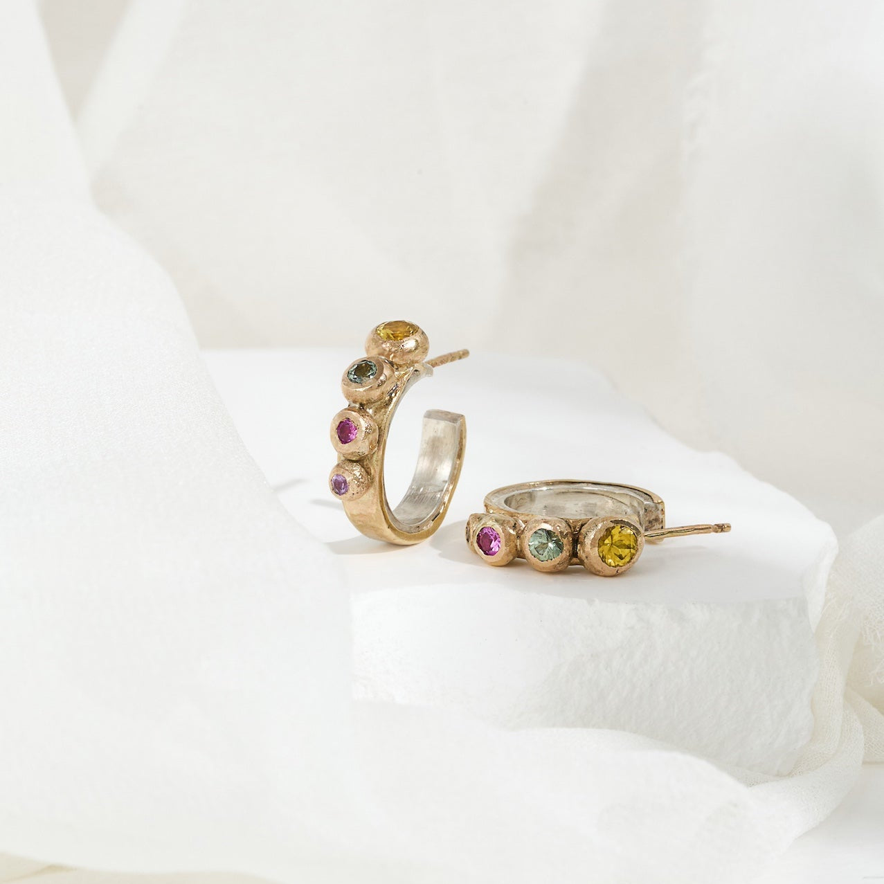 A pair of gold-colored hoop earrings with a gradient of pink, yellow, green, and purple sapphires set in them, displayed on a white fabric surface.