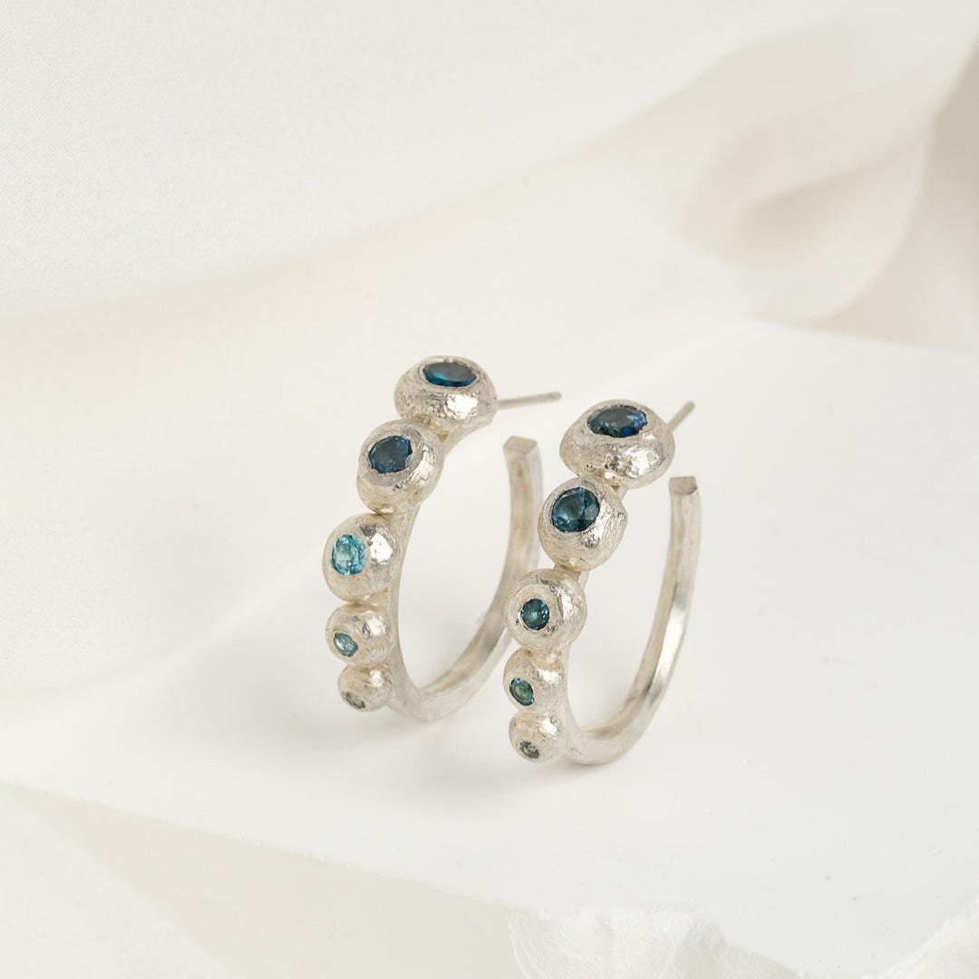 A pair of sterling silver hoop earrings with a gradient of blue topaz stones, arranged from largest to smallest, displayed against a white and pale blue textured background.