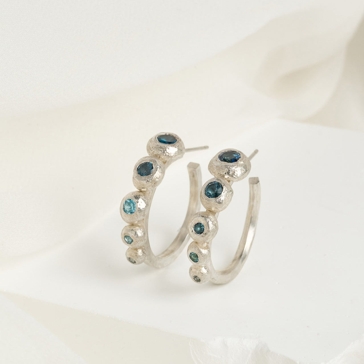 A pair of sterling silver hoop earrings with a gradient of blue topaz stones, arranged from largest to smallest, displayed against a white and pale blue textured background.
