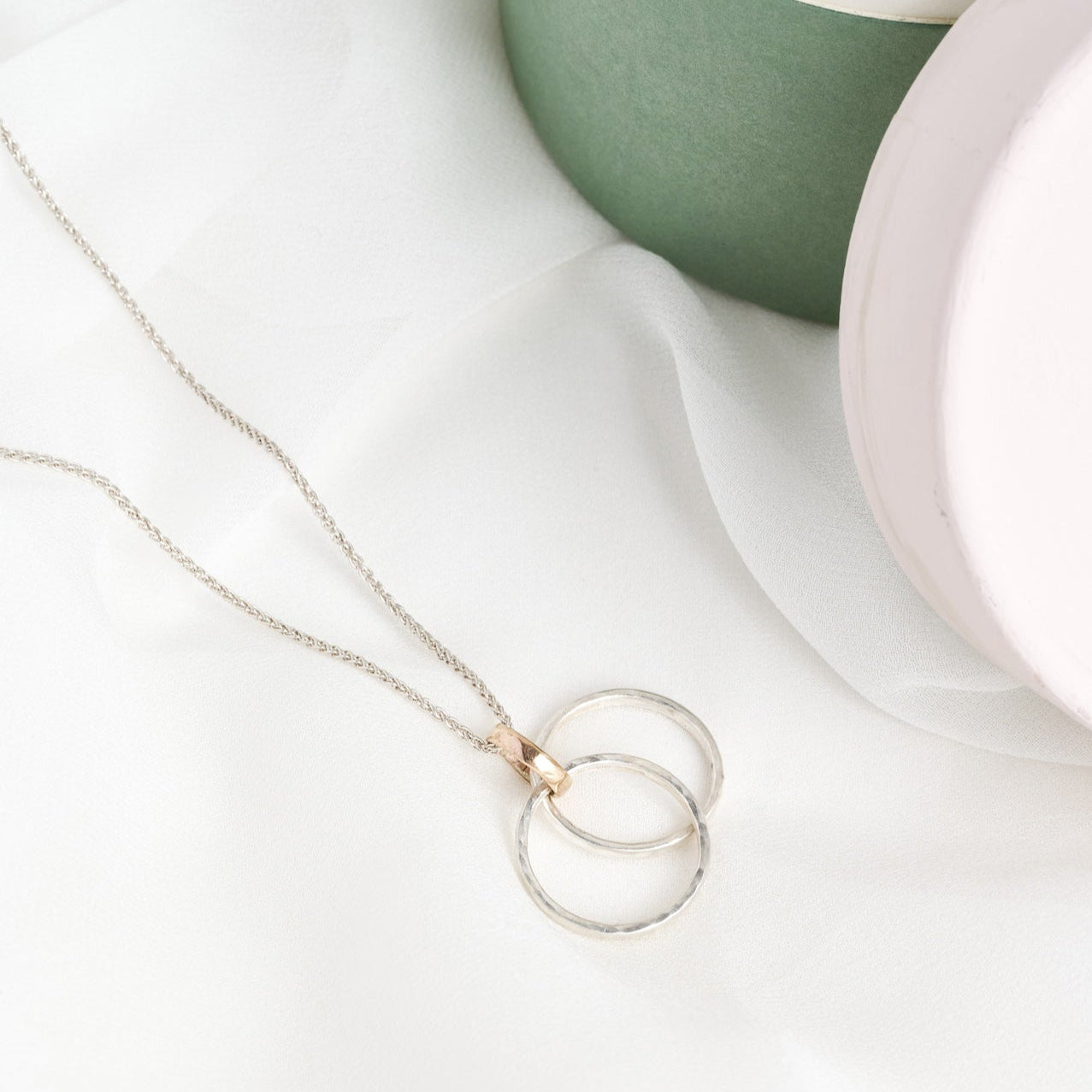 A silver and gold infinity link necklace with a circular pendant lies on a white surface, next to a small potted plant and a round pearl earring.