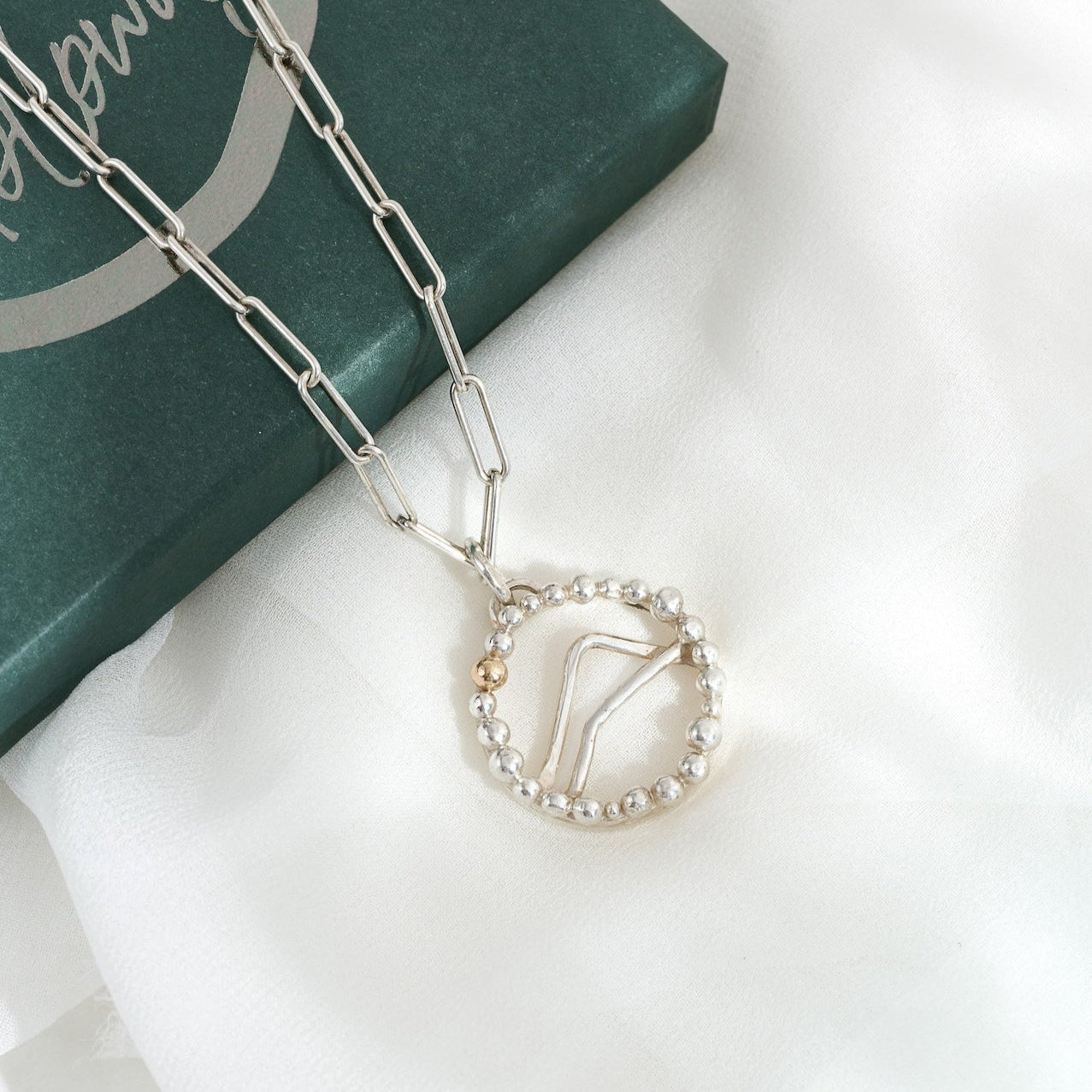 A silver and gold Alpine pendant necklace with a mountainous design, displayed on a white fabric surface next to its green packaging.