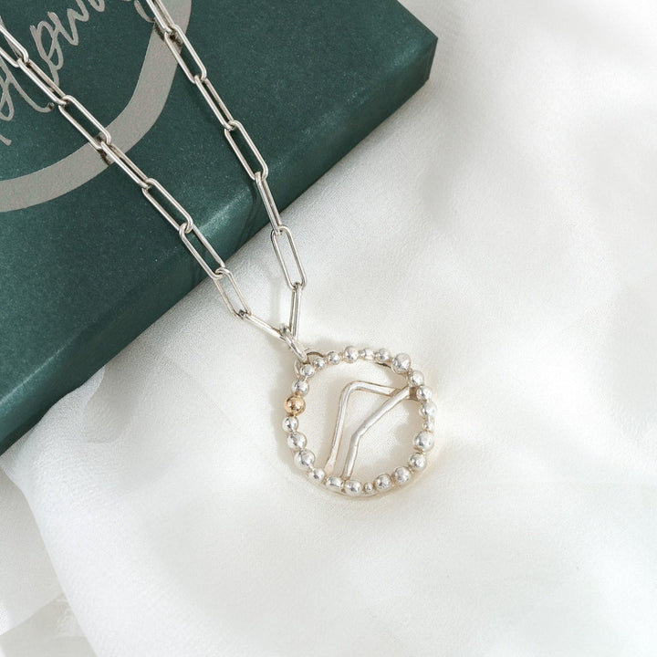 A silver and gold Alpine pendant necklace with a mountainous design, displayed on a white fabric surface next to its green packaging.