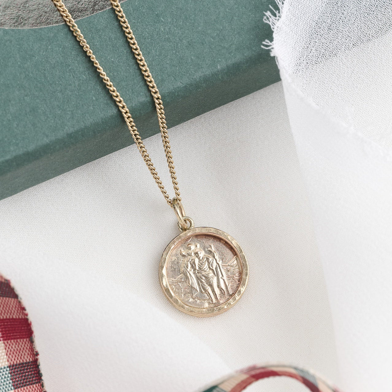A 9ct yellow gold St. Christopher pendant necklace with a curb chain, displayed in front of a box and a plaid fabric.