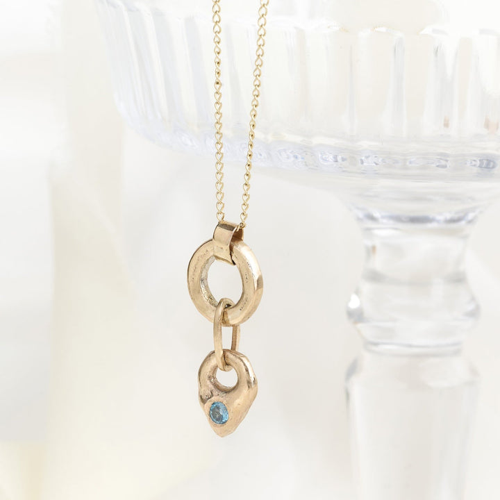 A gold and aquamarine drop necklace with a teardrop-shaped pendant and a chunky gold circle, displayed on a glass stand with a blurred background.