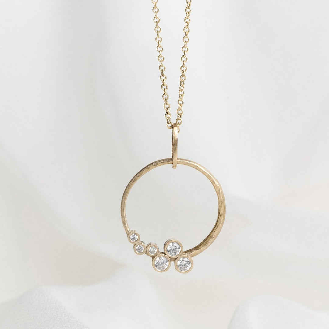 A gold necklace with a circle pendant featuring scattered diamonds, displayed on a white fabric background.
