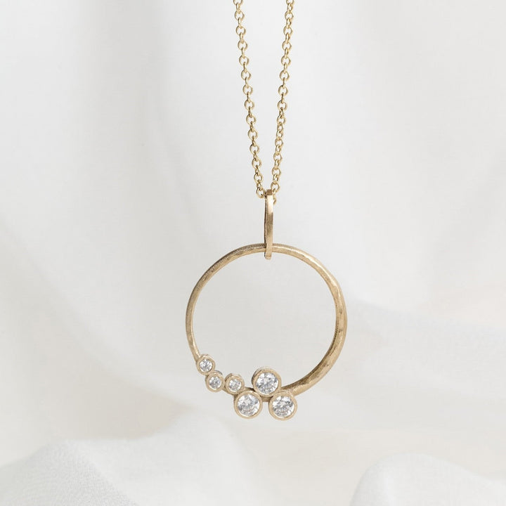 A gold necklace with a circle pendant featuring scattered diamonds, displayed on a white fabric background.