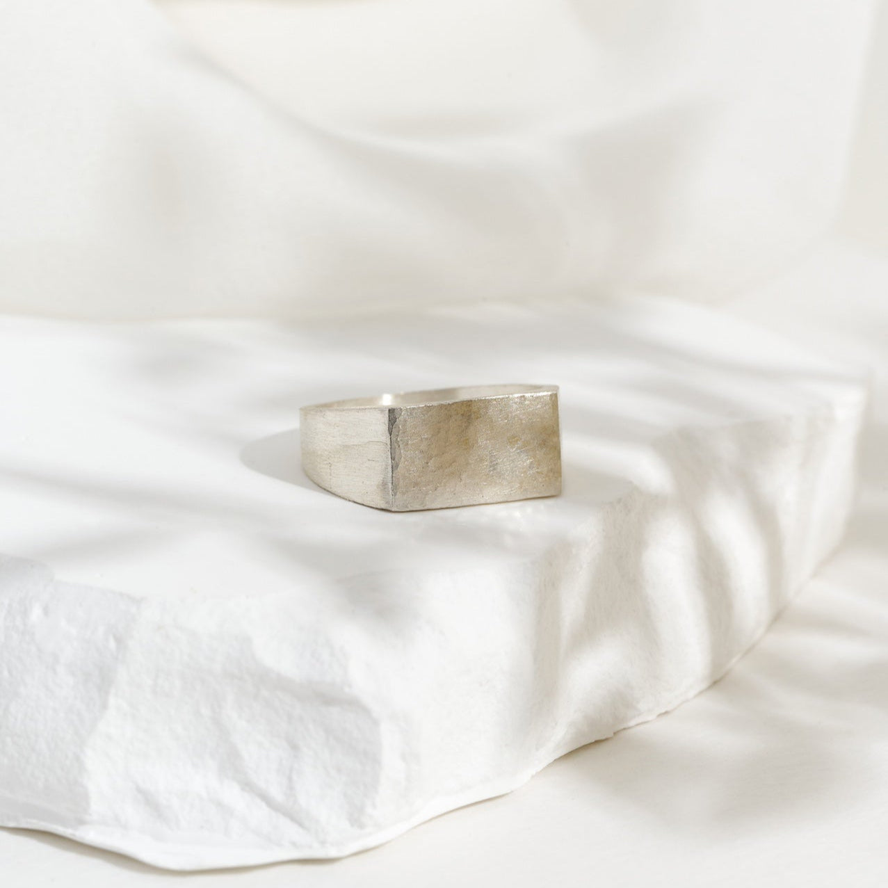 A rectangular silver signet ring placed on a white textured surface, with a blurred background.