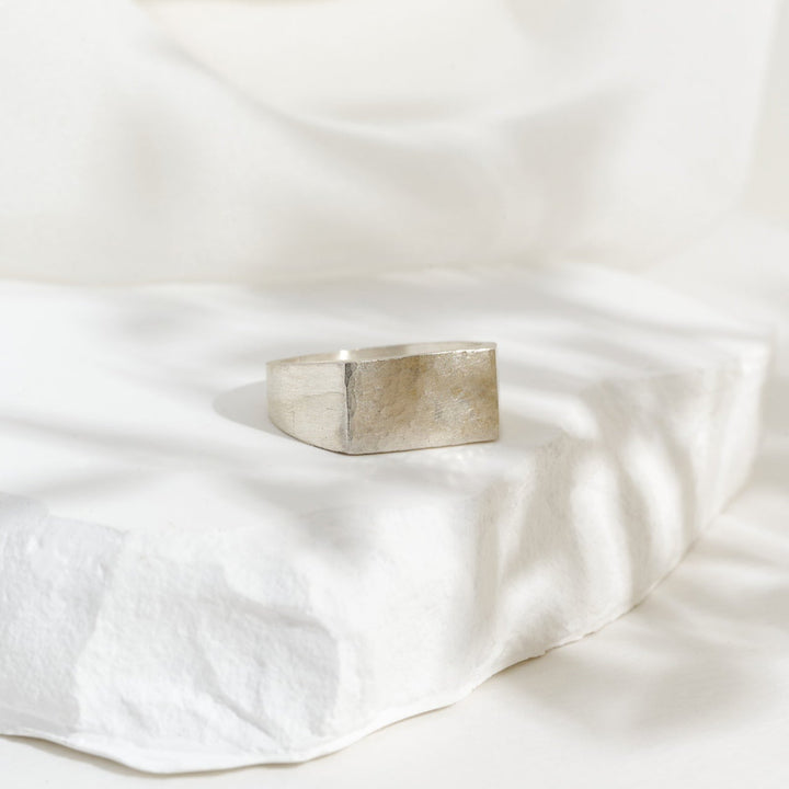 A rectangular silver signet ring placed on a white textured surface, with a blurred background.